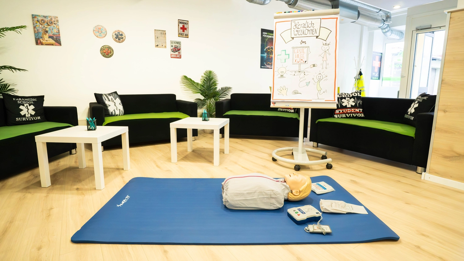 Fahrschule Fahrwerk Bayer GmbH - Führerscheinausbildung - Ein Fahrschulraum in einem Innenraum mit grünen Sofas, einer Tafel und Schulungsmaterialien auf einer blauen Matte auf dem Boden.