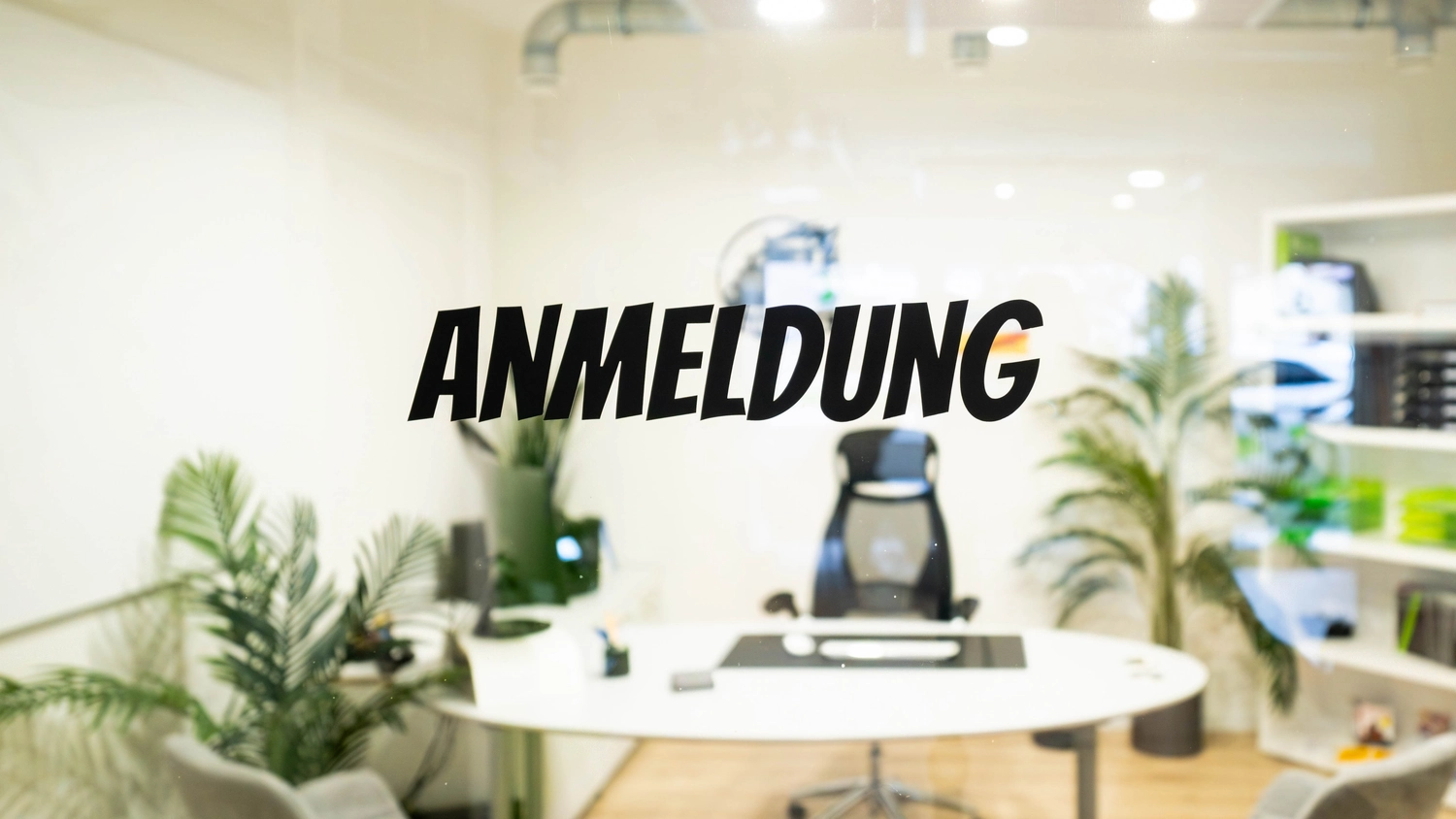 Fahrschule Fahrwerk Bayer GmbH - Führerscheinausbildung - Ein modernes Bürointerieur mit weißem Schreibtisch, schwarzem Bürostuhl, drei grauen Besucherstühlen, mehreren großen Grünpflanzen, einem weißen Bücherregal mit grünen und schwarzen Ordnern und einem schwarzen Logo an der Wand mit einer LKW-Grafik und dem Schriftzug „Fahrwerk“. Der Boden ist mit hellem Laminat ausgelegt.