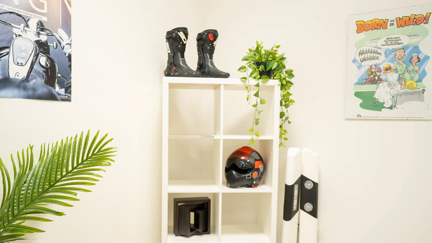 Fahrschule Fahrwerk Bayer GmbH - Führerscheinausbildung - Ein weißes Regal, in dem Motorradbekleidung, darunter ein Helm und Stiefel, ausgestellt ist, steht neben einem Roller.
